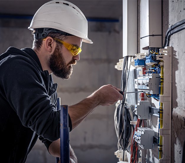 A male electrician works in a switchboard with an electrical connecting cable, connects the equipment with tools.