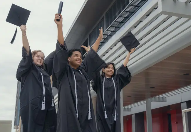 Three students outside in regalia with their hands in the air.